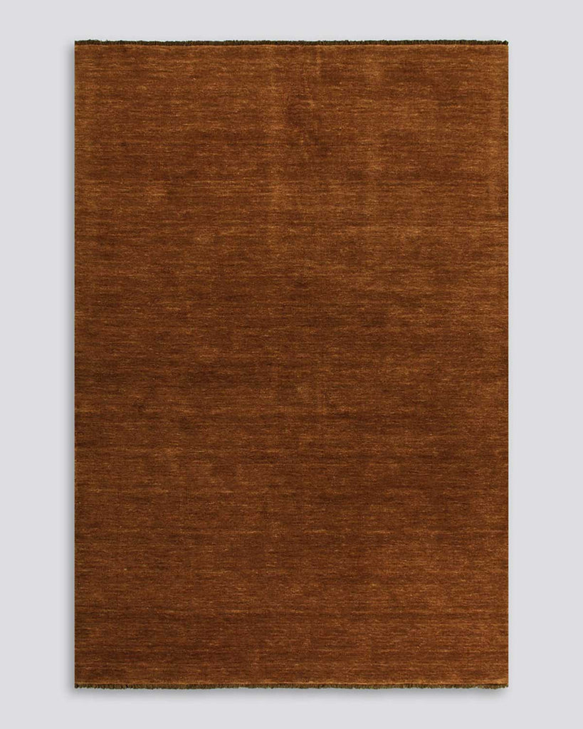 A full image of a warm red-brown floor rug with fringed edges in 100% wool. 