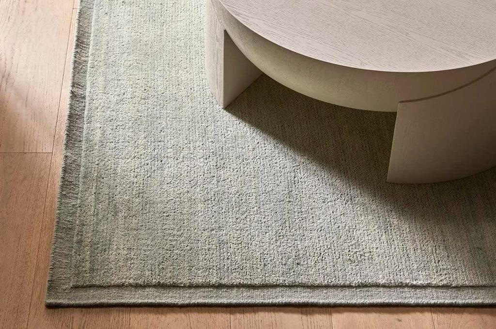 A handmade, 100% wool floor rug situated under a table by Soren Liv NZ and Globe West, in subtle shades of light blue and green, shades of seafoam. It has an indented border, giving interest and sophistication to the rug. 