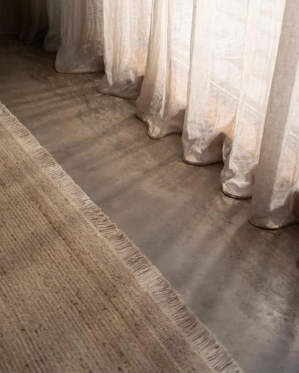 Weave Home Anton wool rug in Sand on a concrete floor beside linen curtains