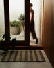 Weave Home Arden jute doormat in Coal at a home entryway, with a person walking through the doorway behind it.