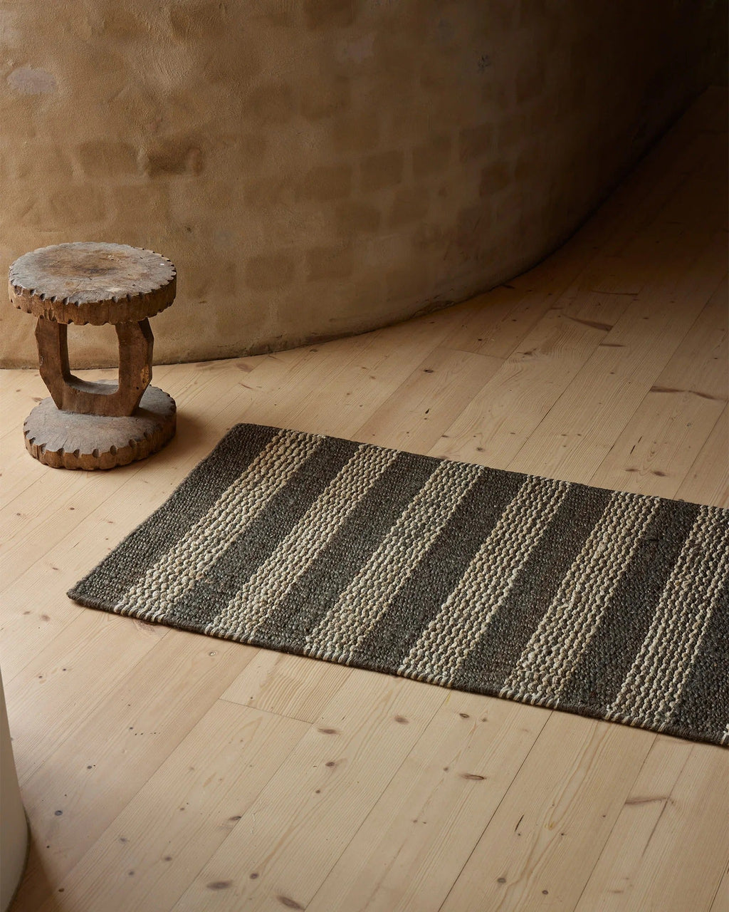 Weave Home Arden jute doormat in Coal styled on a timber floor next to a carved wooden stool.