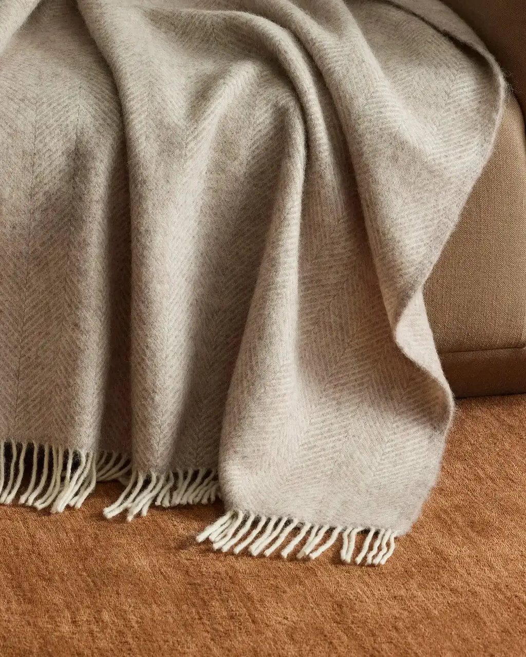 A light, dusty pink with off-white wool herringbone throw blanket, draped on a couch displaying fringe