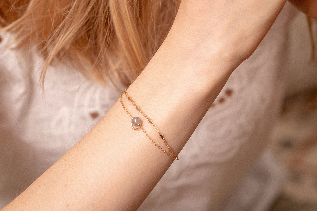A woman wearing a delicate gold chain link bracelet with a grey-green labradorite crystal in the centre. 