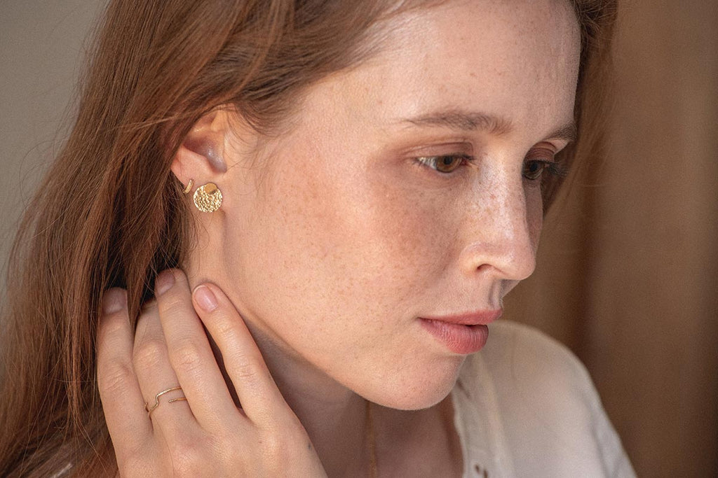 A woman wearing round stud earrings. The hammered jacket of the stud sits in front of her earlobe, catching the light. 