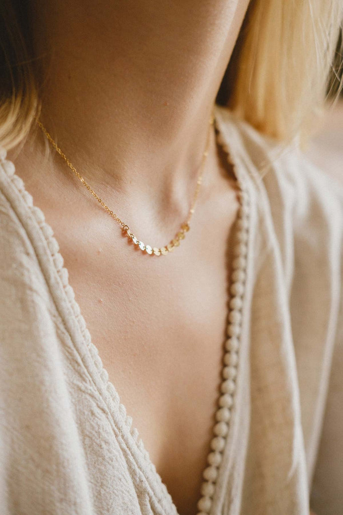 A woman's neck, displaying a delicate gold necklace with small brushed metal gold discs. 