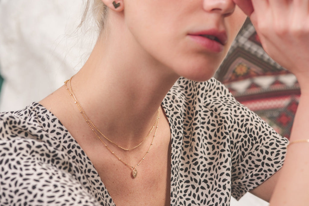 A woman wearing two delicate gold filled necklaces by Zoaje - one is a simple chain with textured flattened sections, and one has a charm in the shape of a sand dollar. 