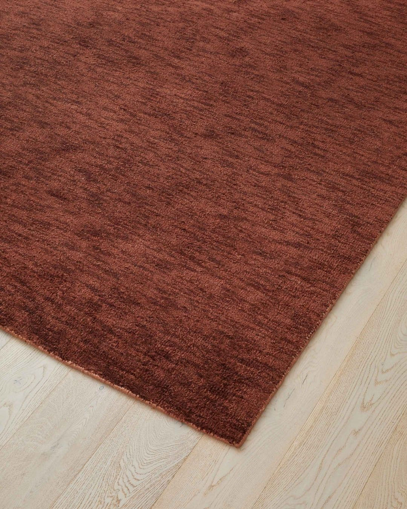 A corner of the Almonte floor rug by Weave Home, showing its earthy red tone and texture