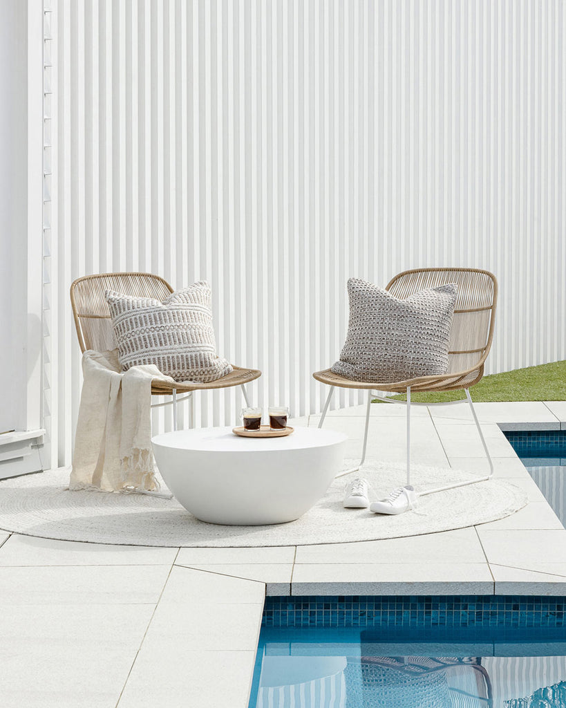 Two chairs and a table arranged outside by a swimming pool on a sunny day. They are sitting on top of a white-toned rug by Baya NZ. 