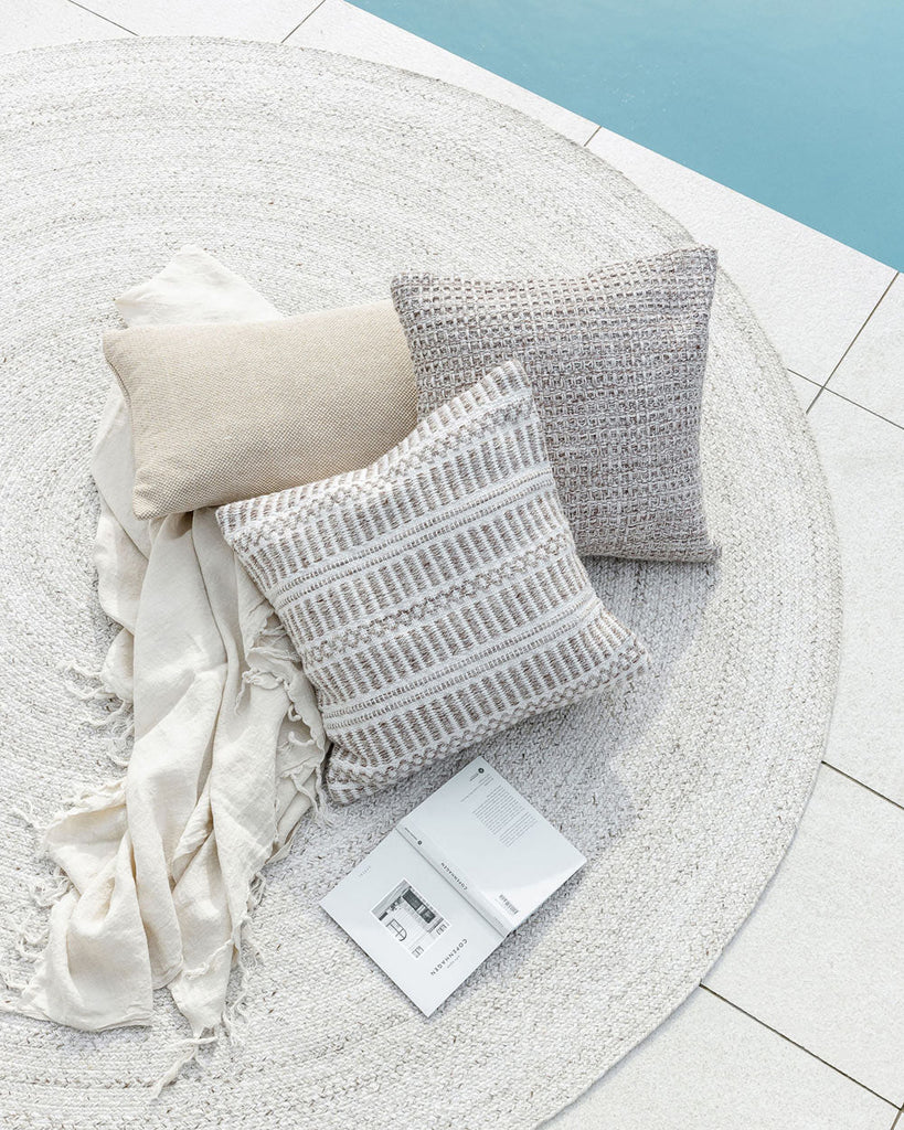 A white outdoor rug by Baya NZ, sitting by a swimming pool with cushions and a throw. 